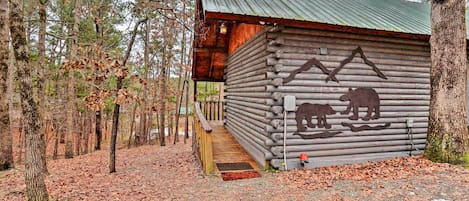 A rustic log cabin with a wooden staircase leading to a porch, surrounded by a forest.