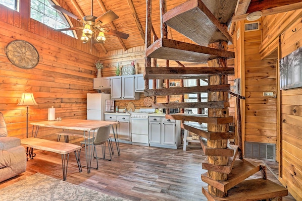 A rustic cabin interior with a wooden dining table, chairs, a kitchen area, and a spiral staircase. - Broken Bow Log Cabin w/ Fire Pit, Pond, & Deck! (Broken Bow)