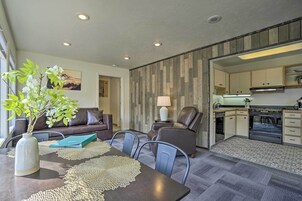 Una sala moderna con un sofá, sillones, una mesa de comedor con un jarrón y individuales, una cocina con armarios y electrodomésticos, y un piso con diseño.