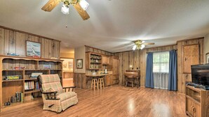 A spacious living room with a wooden floor, a plaid armchair, a dining area with stools, a kitchen with wooden cabinets, and a ceiling fan. - Cozy Charlotte Retreat w/ Yard ~ 5 Mi to Dtwn (Charlotte)