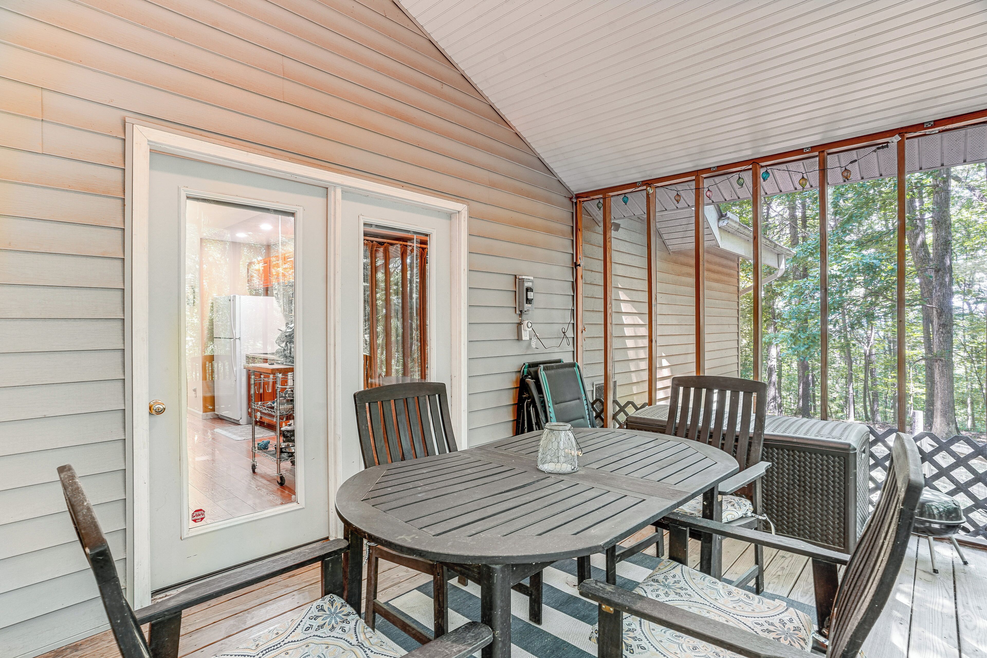 A covered patio with a table and chairs, a sliding glass door leading to a kitchen, and a view of trees through the glass.