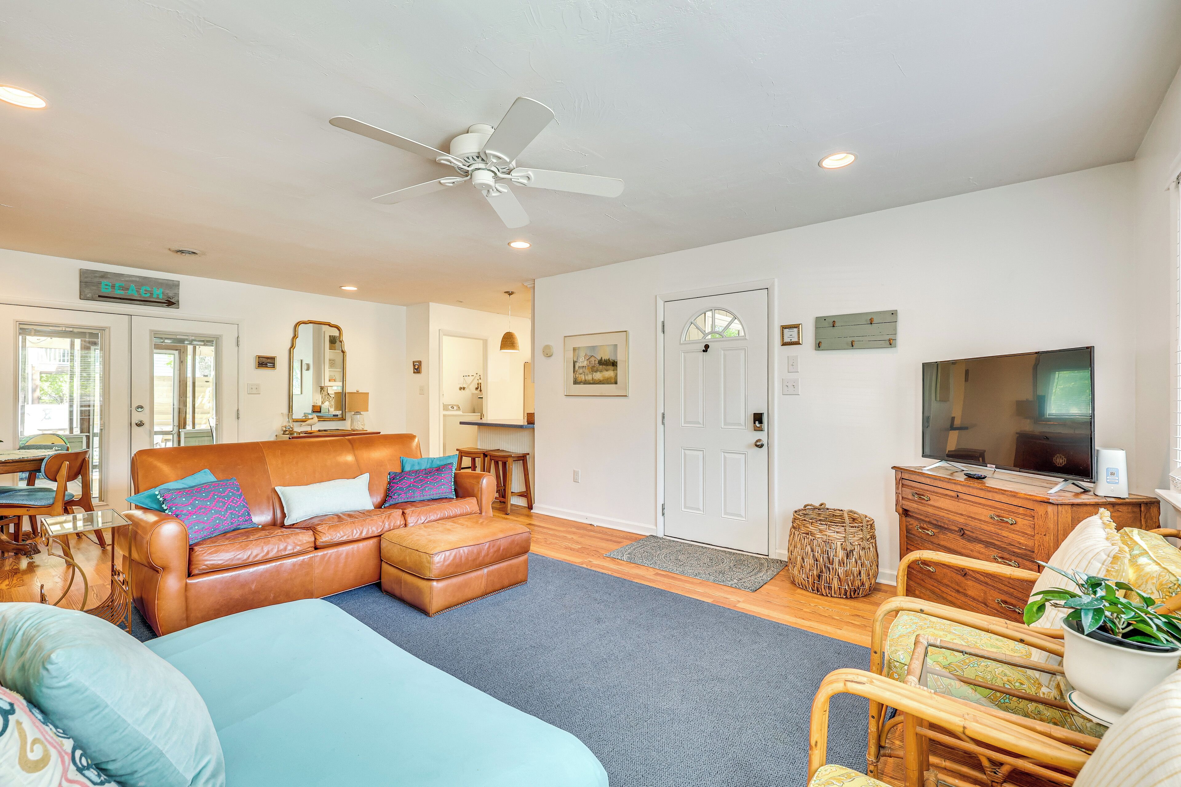 A spacious living room with a brown leather sofa, a blue couch, and a wooden chair. There is a television on a wooden cabinet, a ceiling fan, and a door labeled "STAGE".