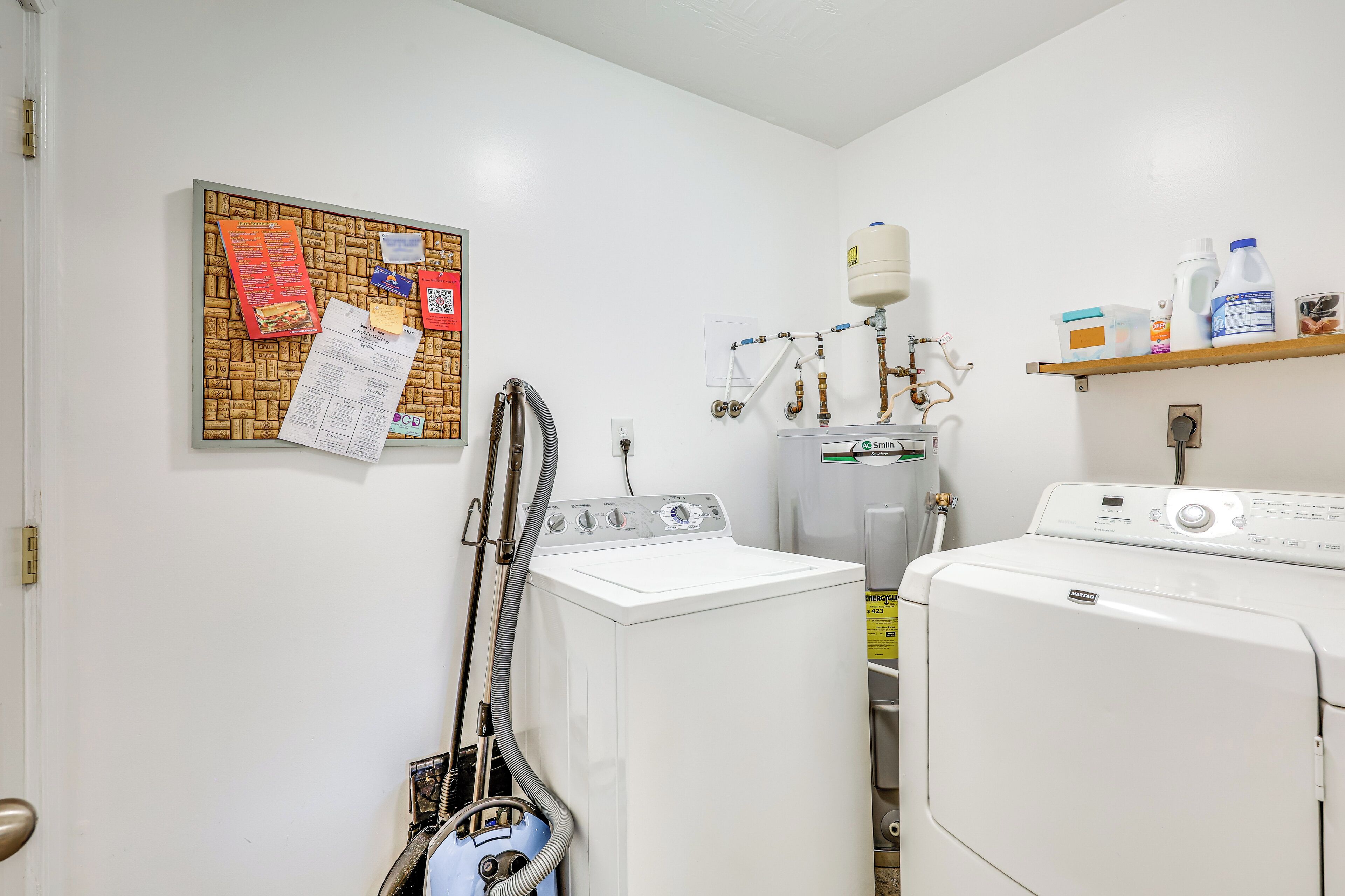 A laundry room with two washing machines, a water heater, and a shelf with cleaning supplies.