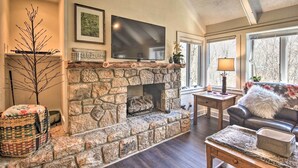 A stone fireplace with a flat-screen TV above it, a cozy seating area with a sofa and a coffee table, and a window providing natural light. - Steps to Ski Resort: Sugar Mountain Condo! (Banner Elk)