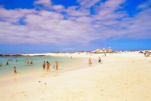 A beach with people swimming and walking on the sand, and a building in the background.