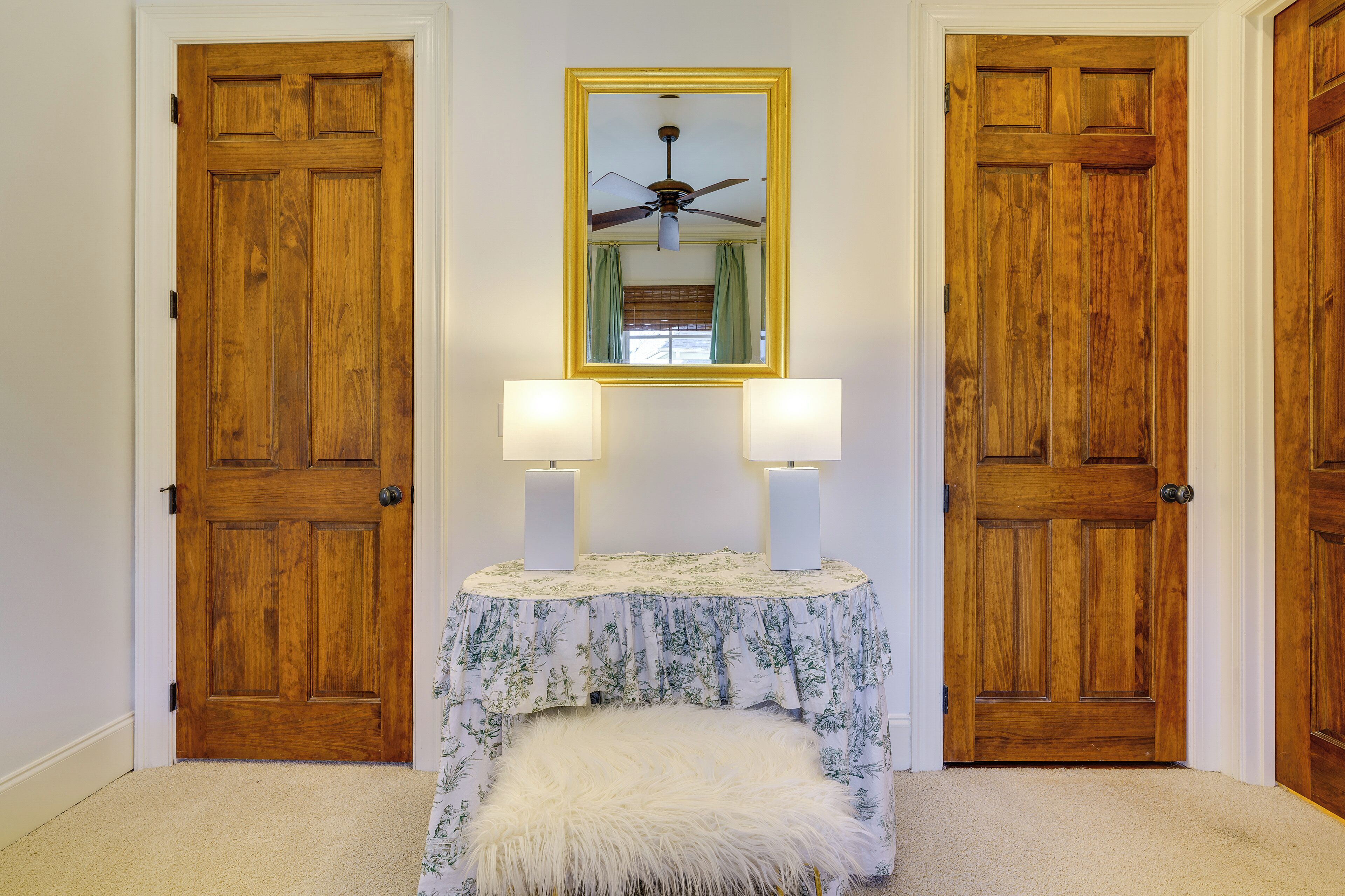 Una habitación con dos puertas de madera, un espejo con marco amarillo, un ventilador de techo y una pequeña mesa con dos lámparas.