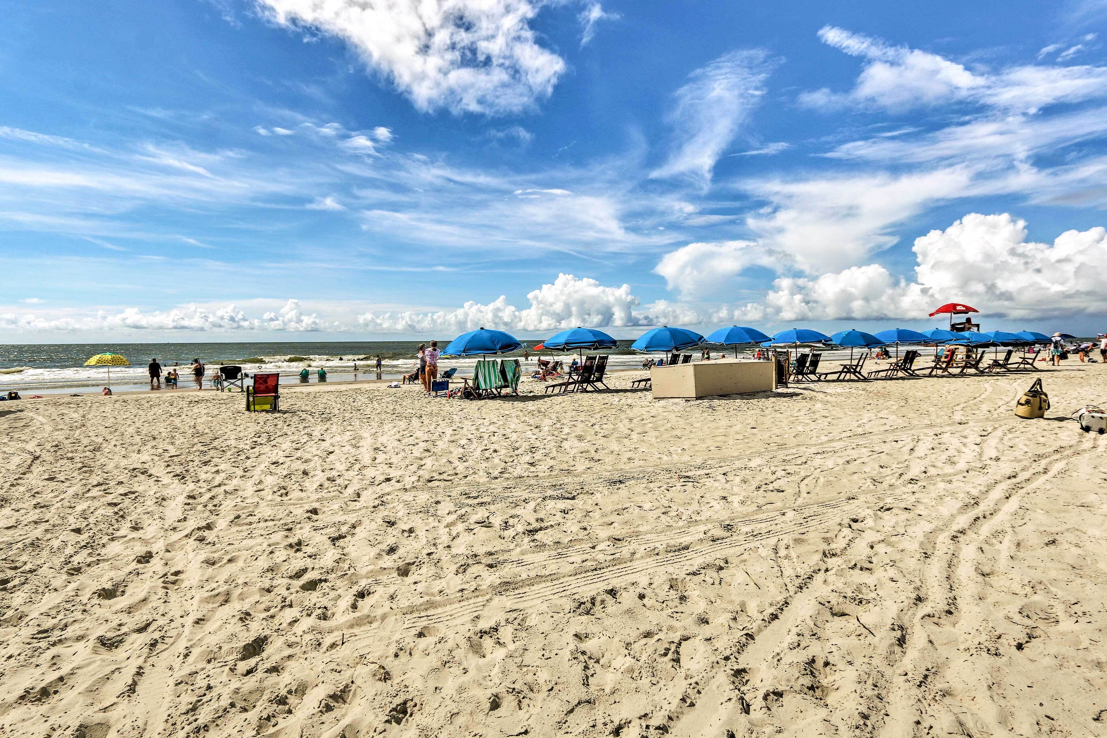A sandy beach with blue umbrellas and beach chairs, a lifeguard tower, and the ocean in the background.