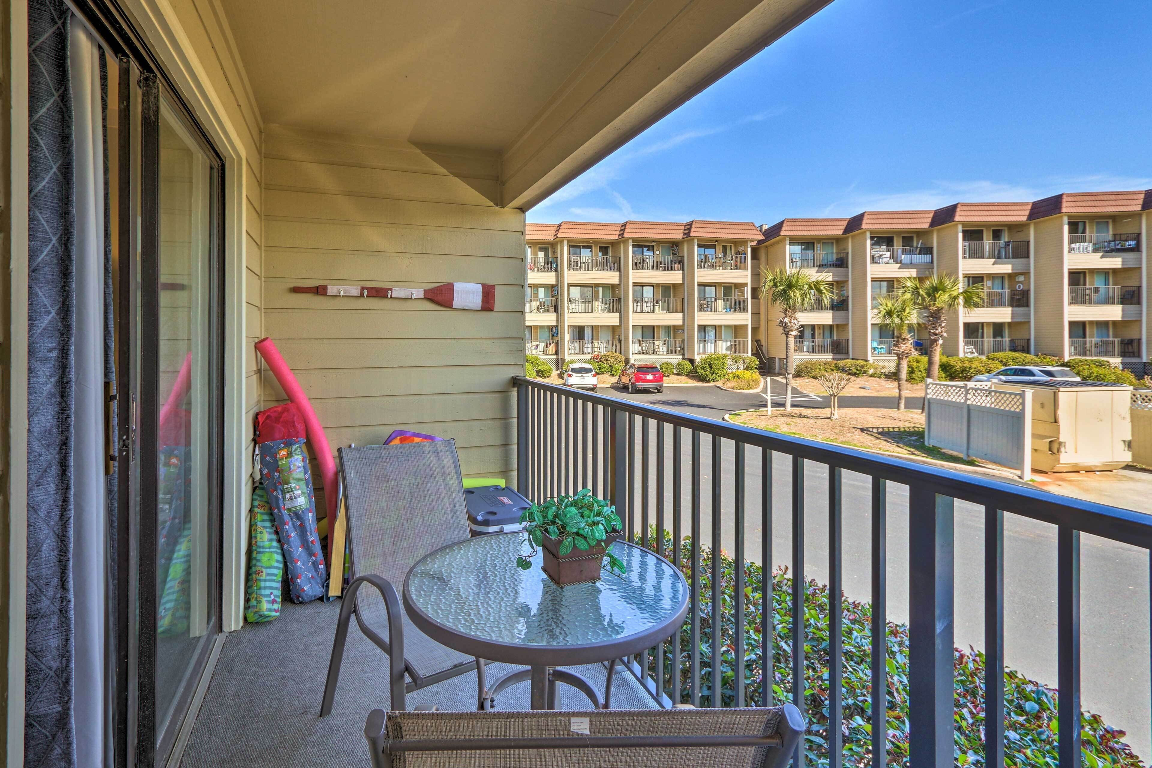 A balcony with a glass table and chairs, a potted plant, and a view of a multi-story building with balconies.