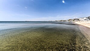 A beach with clear shallow waters, a sandy shore, and a row of buildings in the background. - Westbrook Home w/ Ocean Views - Walk to Beach (Westbrook)