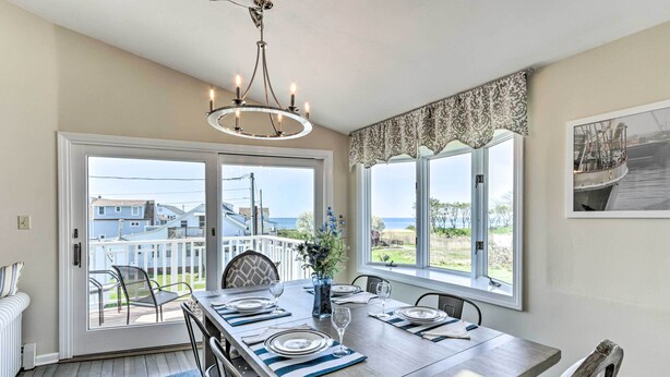 A dining area with a large table set for six, a chandelier, and a view of the ocean through large windows. - Westbrook Home w/ Ocean Views - Walk to Beach (Westbrook)
