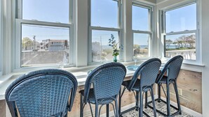 A dining area with wicker chairs and a table, large windows with a view of the sea and buildings, and a vase with flowers on the table. - Westbrook Home w/ Ocean Views - Walk to Beach (Westbrook)
