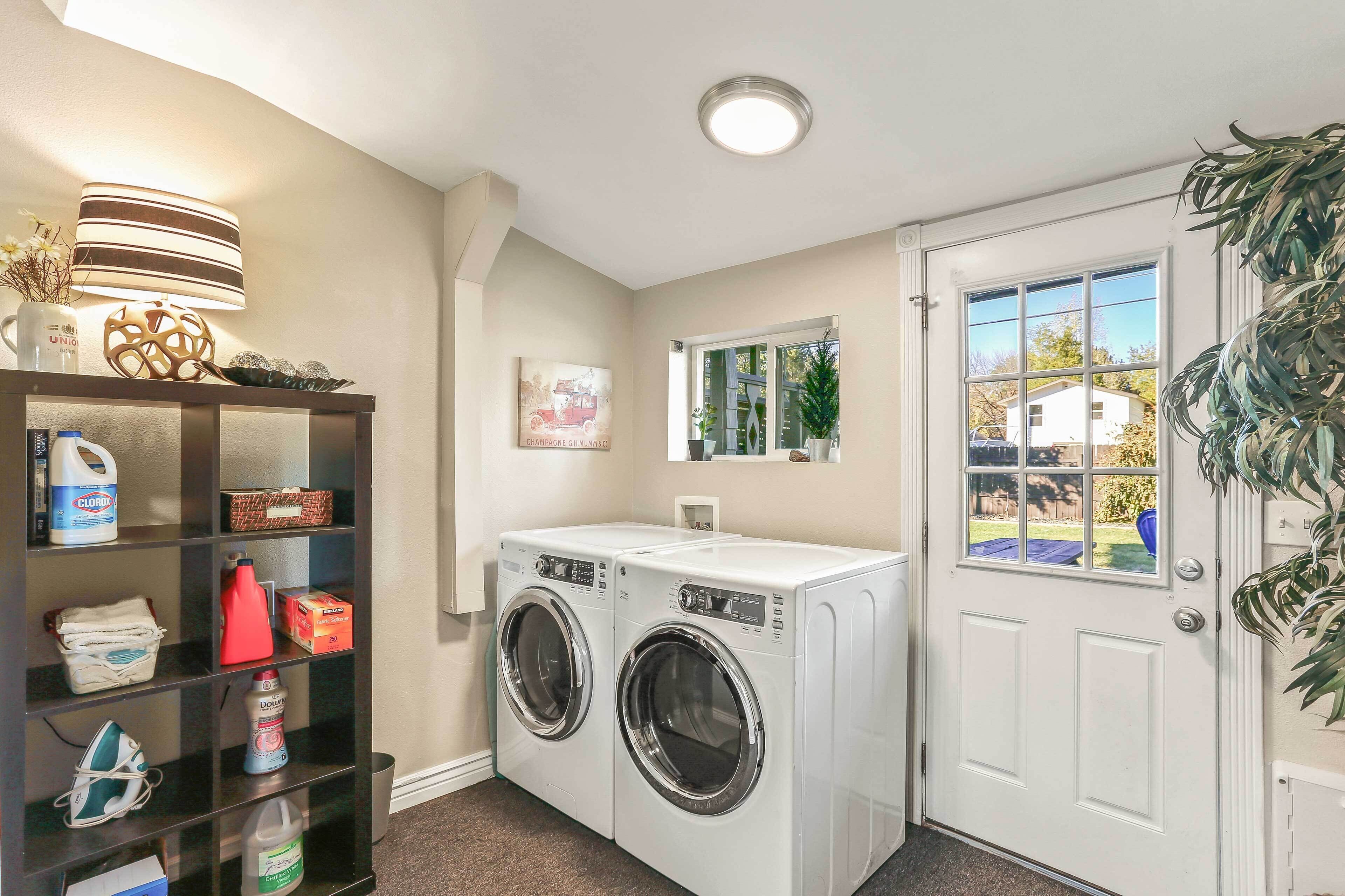 A laundry area with two front-loading washers, a shelf with cleaning supplies, a decorative lamp, and a window with a view of the outside.