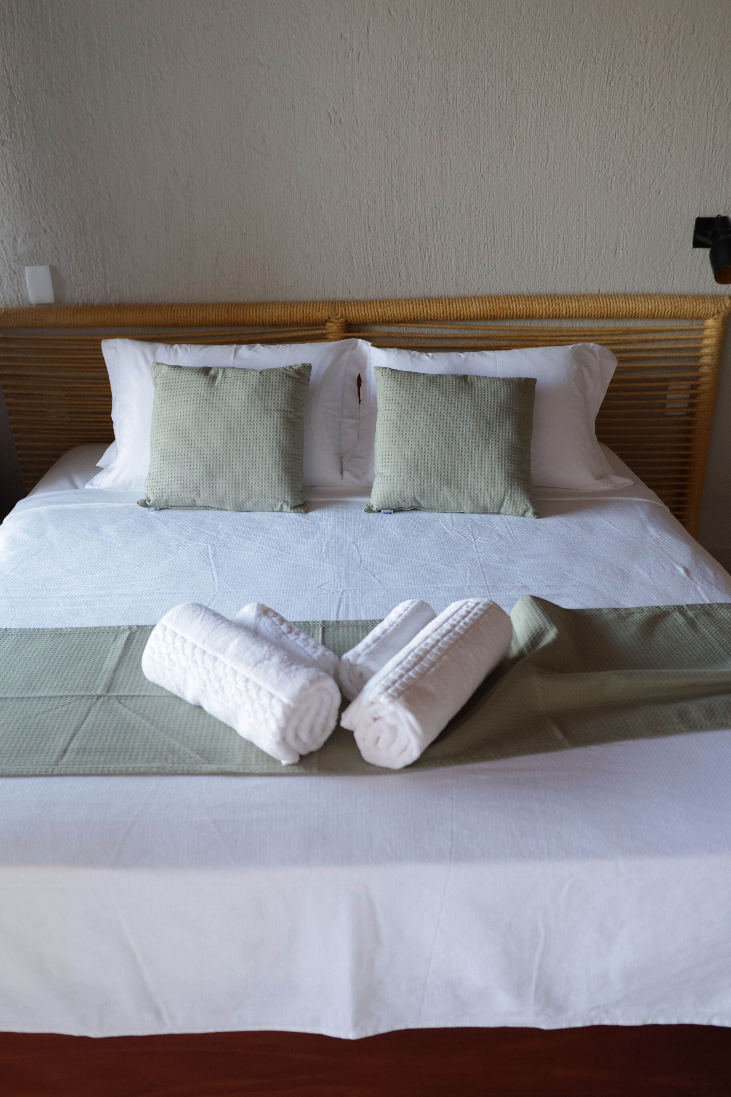 Una cama bien tendida con sábanas blancas, dos almohadas verdes y dos toallas blancas enrolladas.