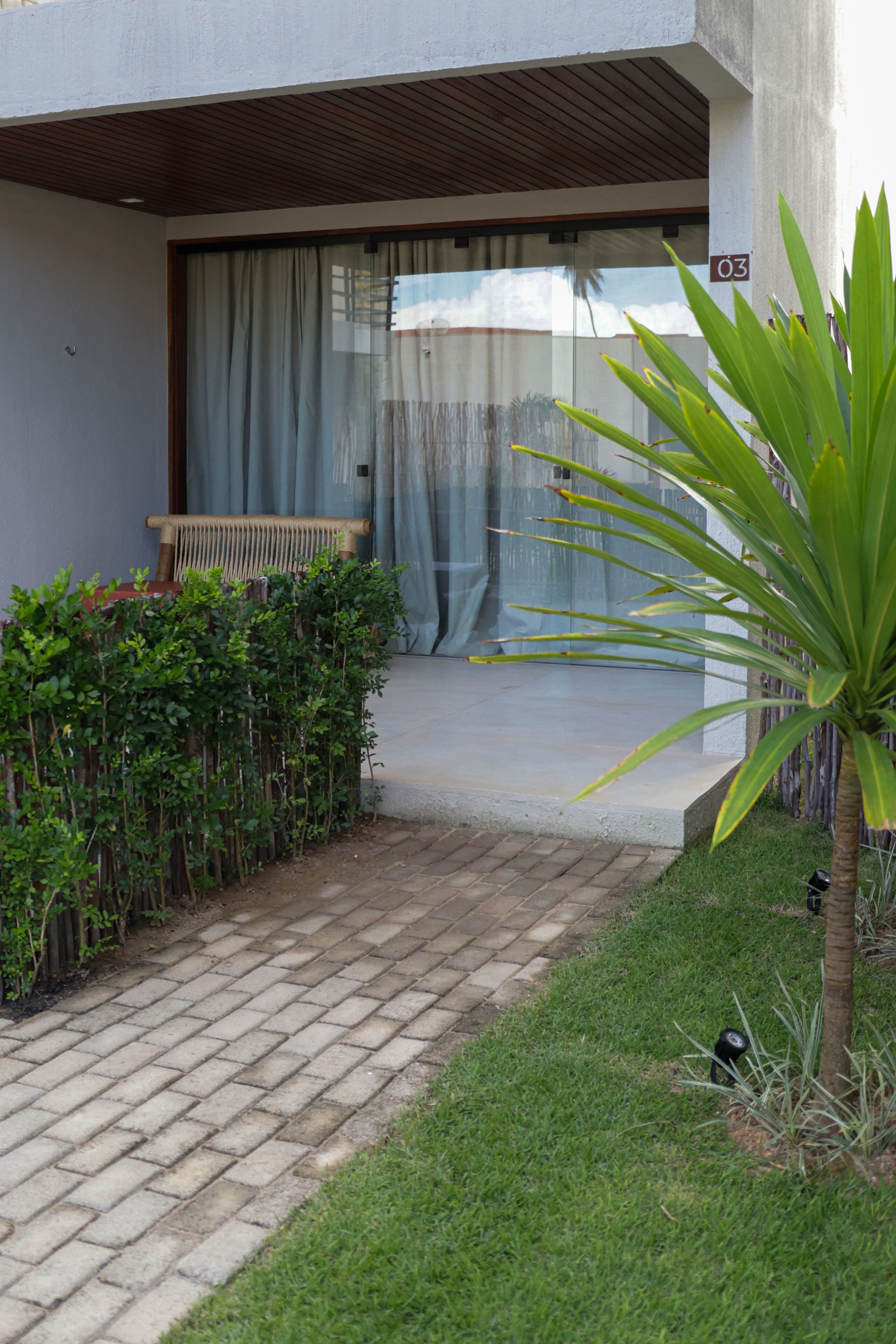 Un camino pavimentado que lleva a la entrada de un edificio, con un banco de madera y una planta.