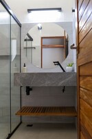 Un baño moderno con mampara de vidrio, espejo redondo, repisa de madera y una pequeña planta en maceta.