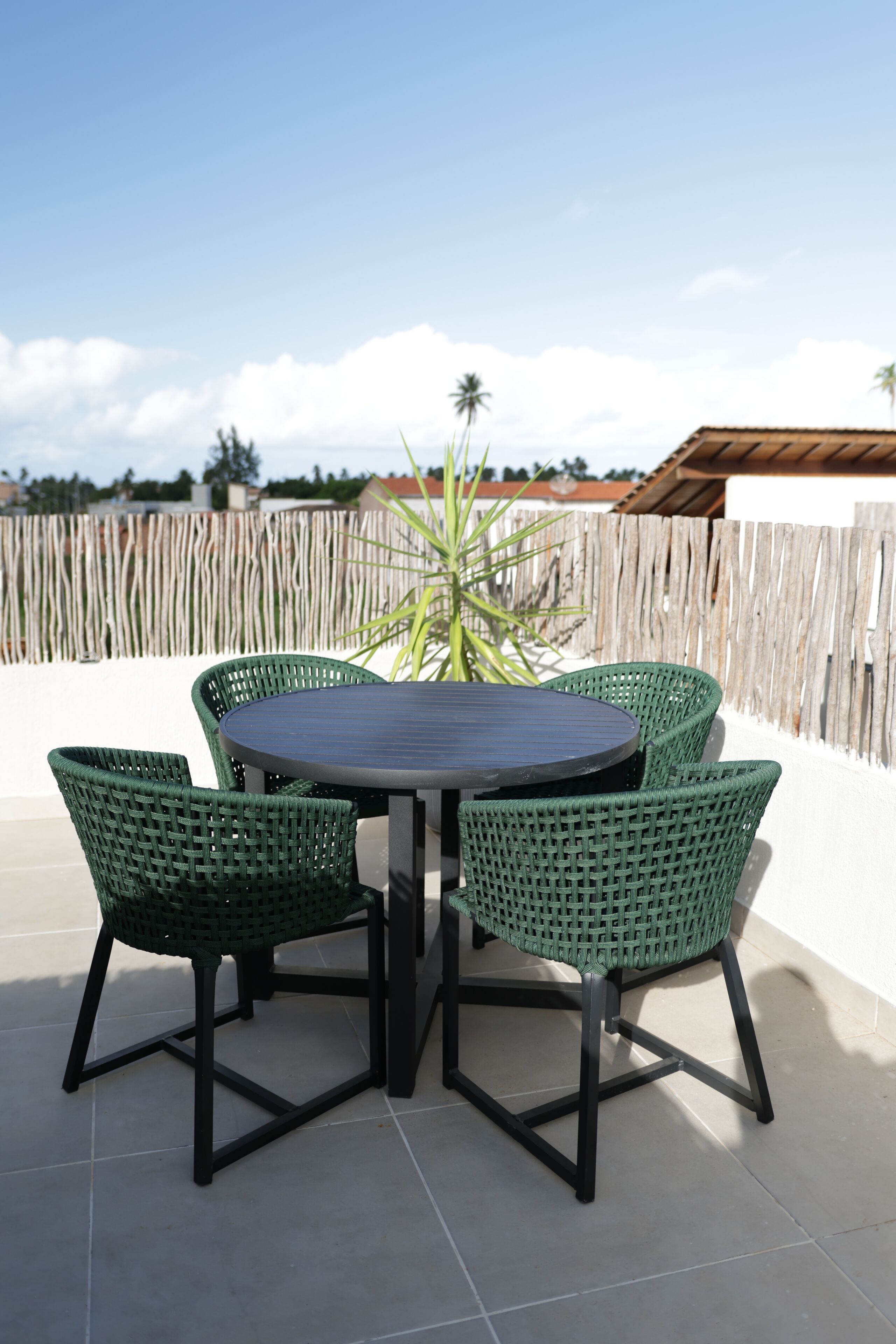 A round table with four green chairs, a wooden fence, and a palm tree in the background.
