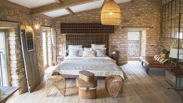 A rustic bedroom with a stone wall, a large bed with multiple pillows, a wooden bench, and wicker furniture.