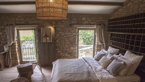 A rustic bedroom with a stone wall, a wooden bed, wicker furniture, and a view through a window.