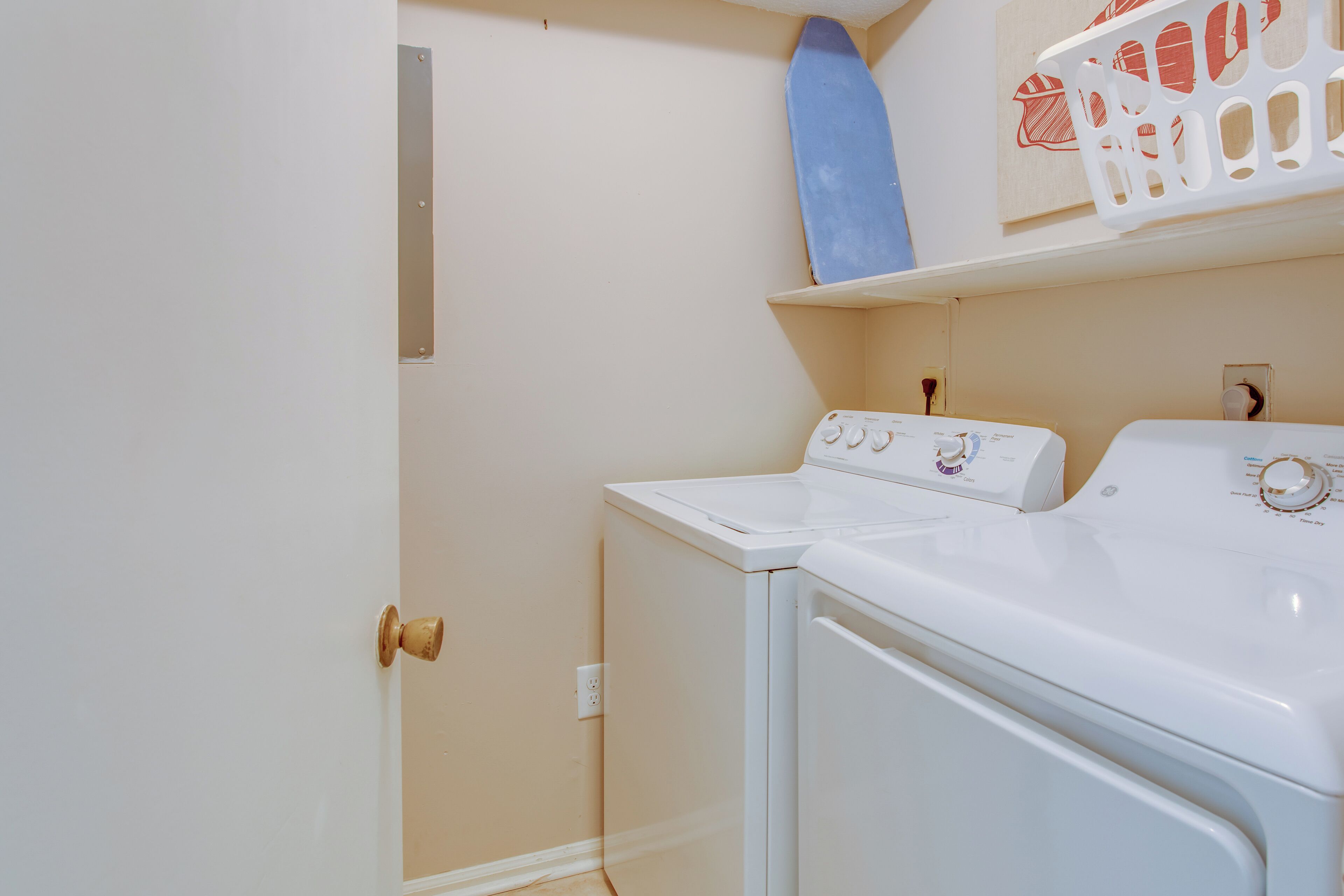 A laundry room with a front-loading washer and dryer, a wooden door, and a shelf above the dryer.