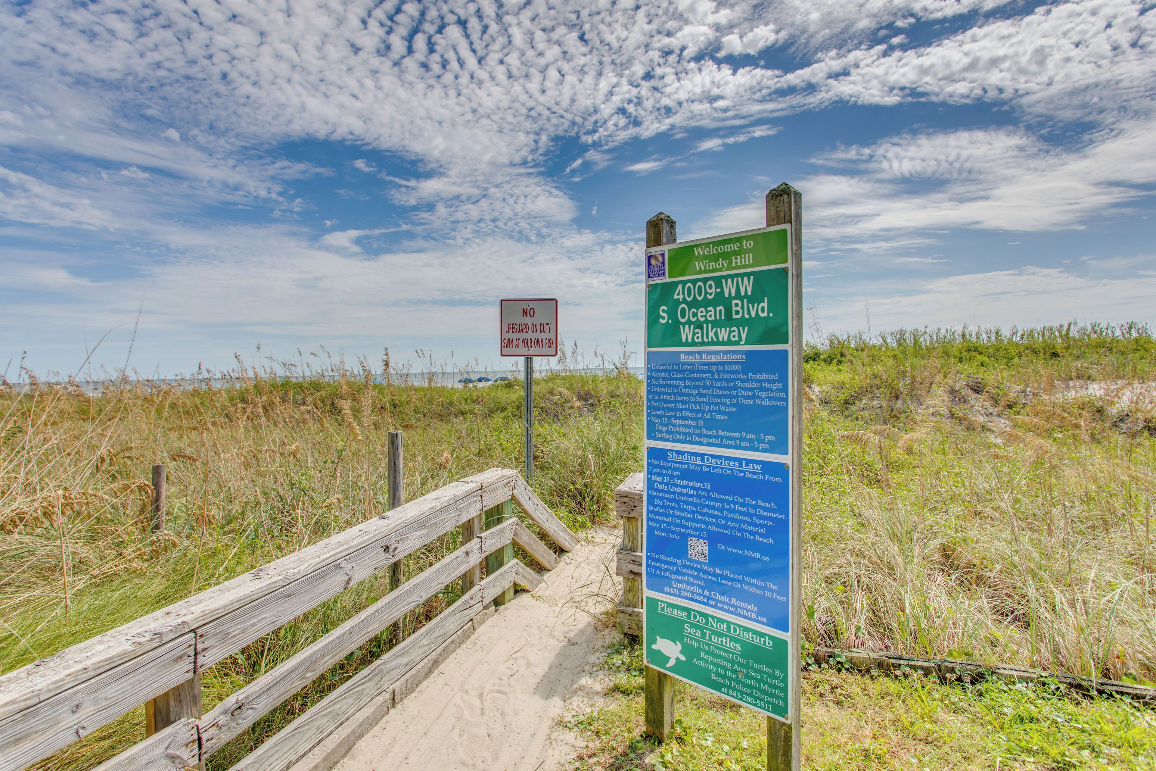 Un malecón de madera que conduce a una playa, con un cartel que indica '4009-WW S. Ocean Blvd Walkway' y 'SIN SOCORRISTAS DE GUARDIA'.