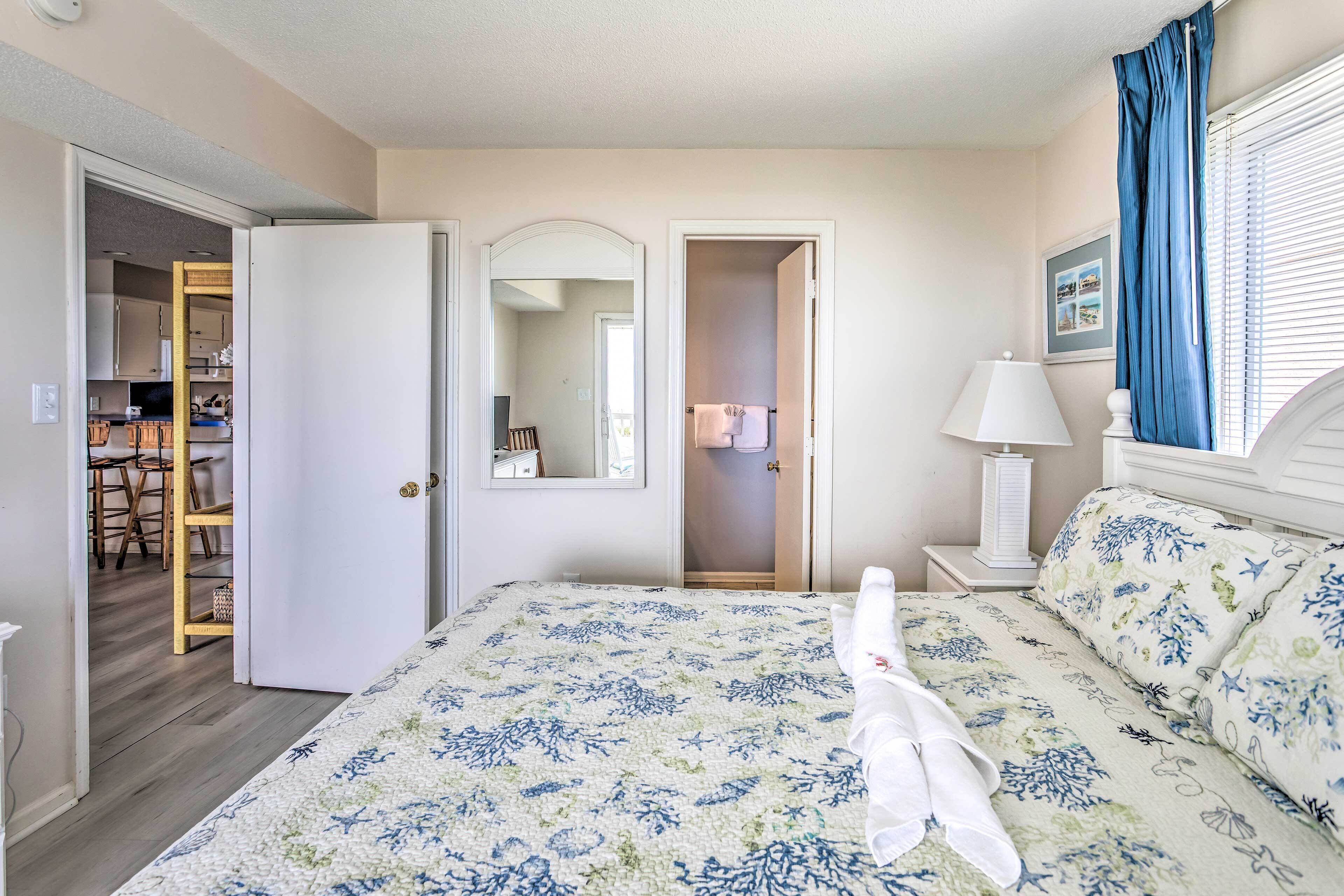 A hotel room with a bed, a nightstand, a lamp, a window with curtains, and a door leading to a bathroom.