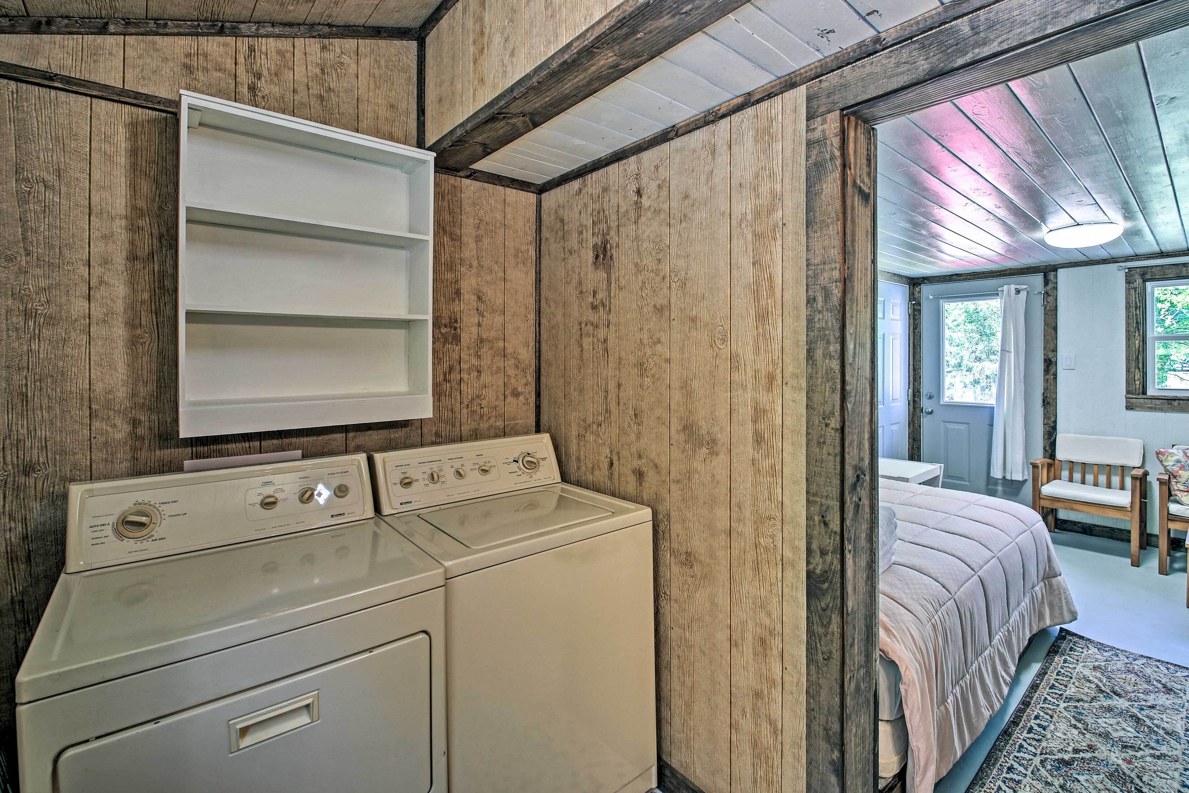 A laundry room with a washer and dryer, wooden walls, and a view into a bedroom with a bed and a window.
