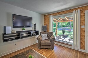Een woonkamer met een flatscreen televisie, een bruine leren fauteuil, een houten vloer en uitzicht op een terras met tuinmeubels.