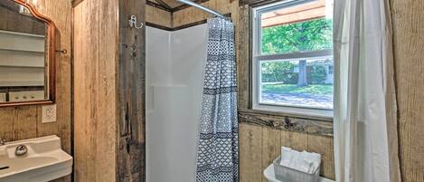 A rustic bathroom with a wooden interior, a shower with patterned curtains, a sink, and a window with a view of greenery.