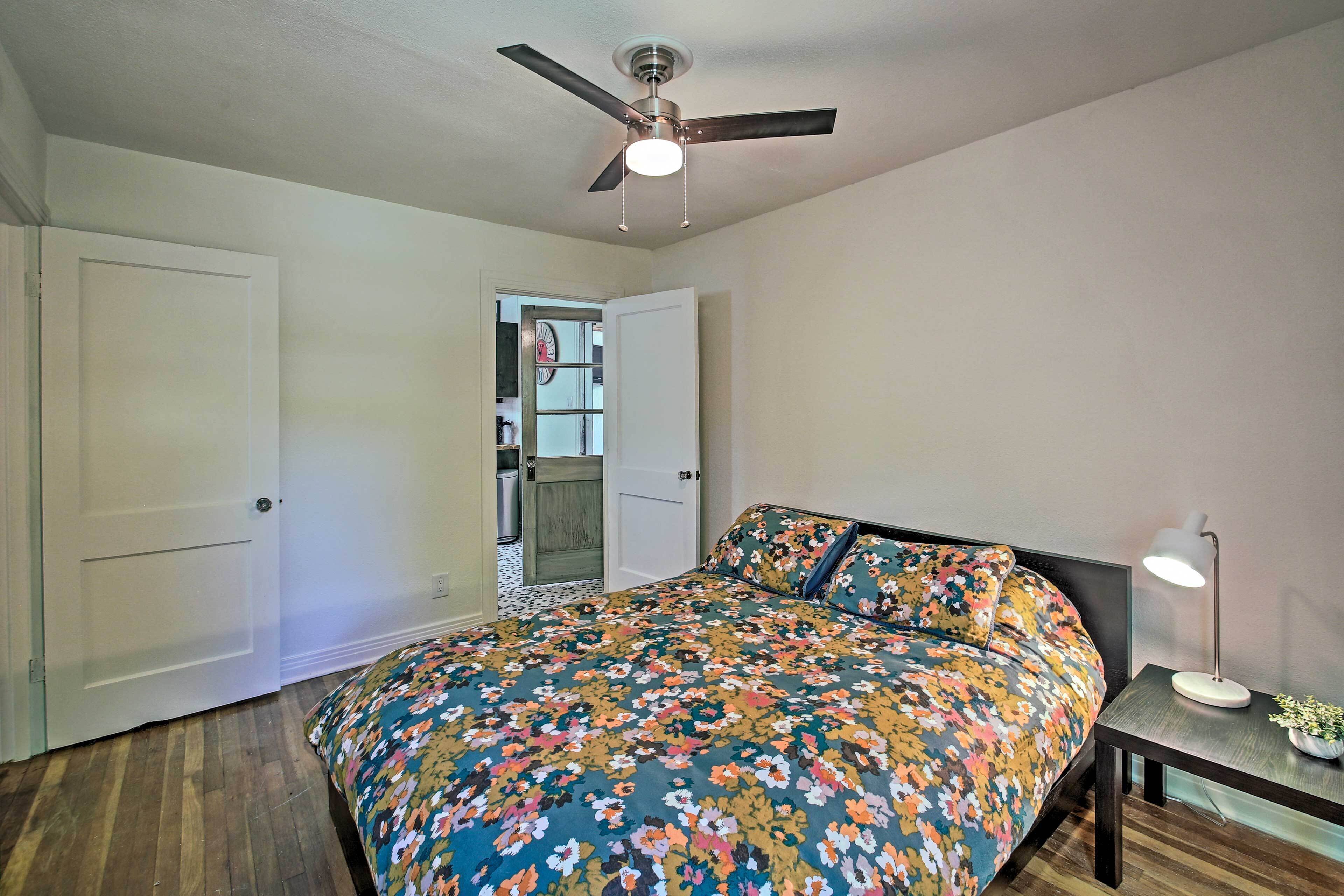 A bedroom with a bed, a ceiling fan, a nightstand with a lamp, and a door leading to another room.