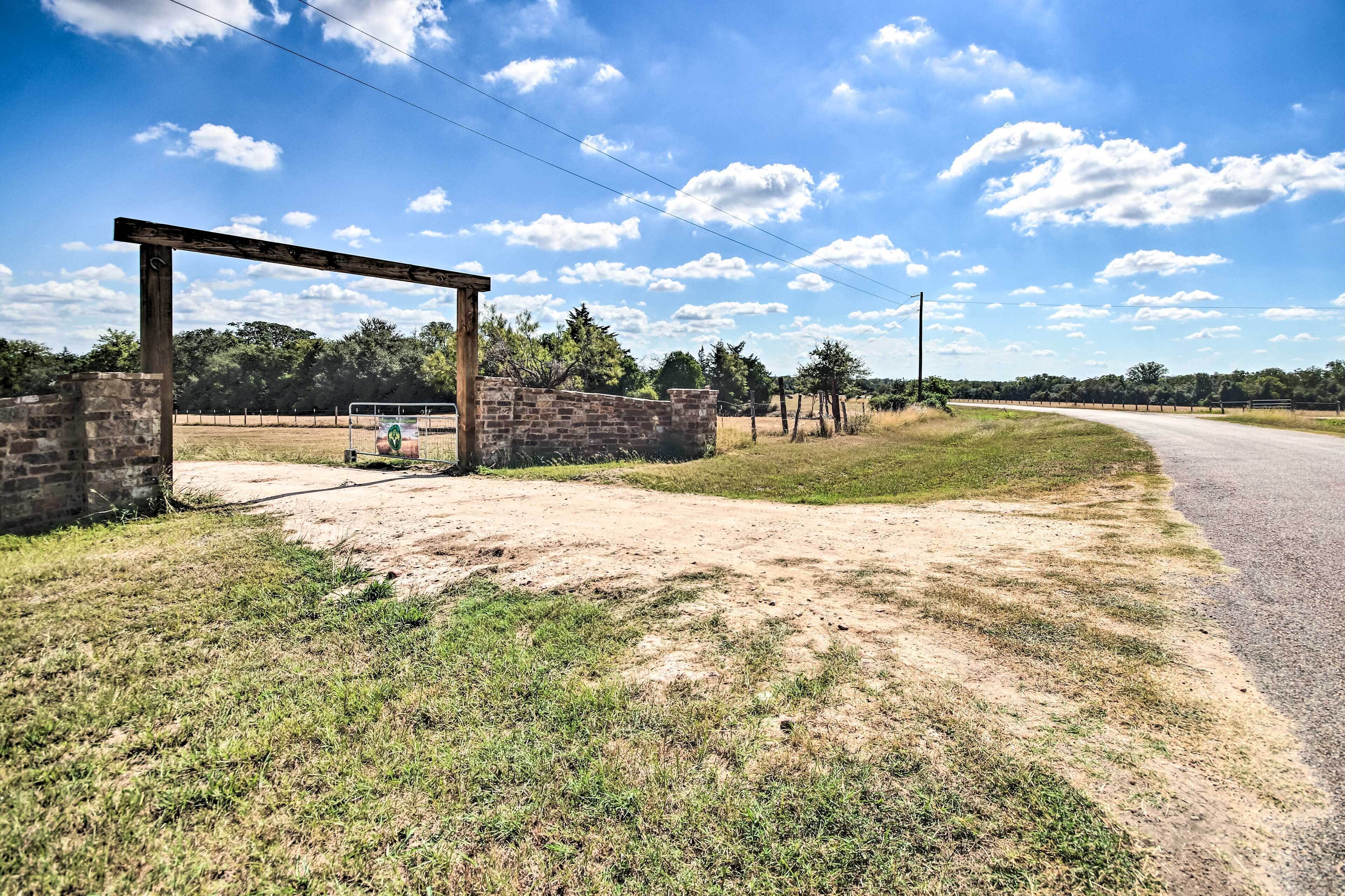 A rural road with a dirt path and a wooden archway leading to a grassy area.