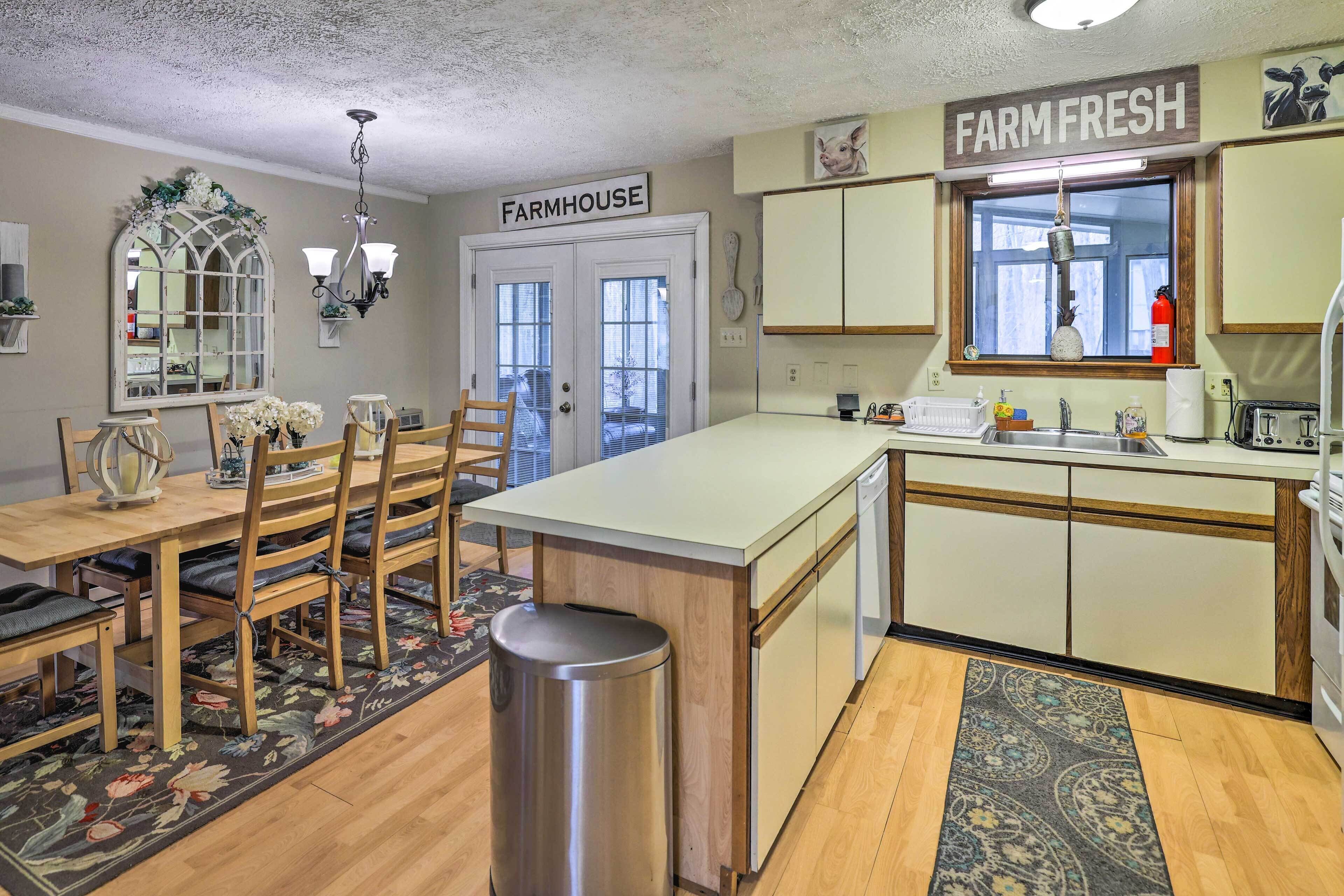 A kitchen with a farmhouse sign, wooden chairs, a dining table, and a small trash bin.
