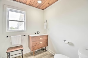 A bathroom with a wooden ceiling, a white sink, a wooden vanity, a white toilet, and a window with a towel hanging on the wall. - Rustic Beachfront Cottage w/ Deck & Boardwalk (Supply)