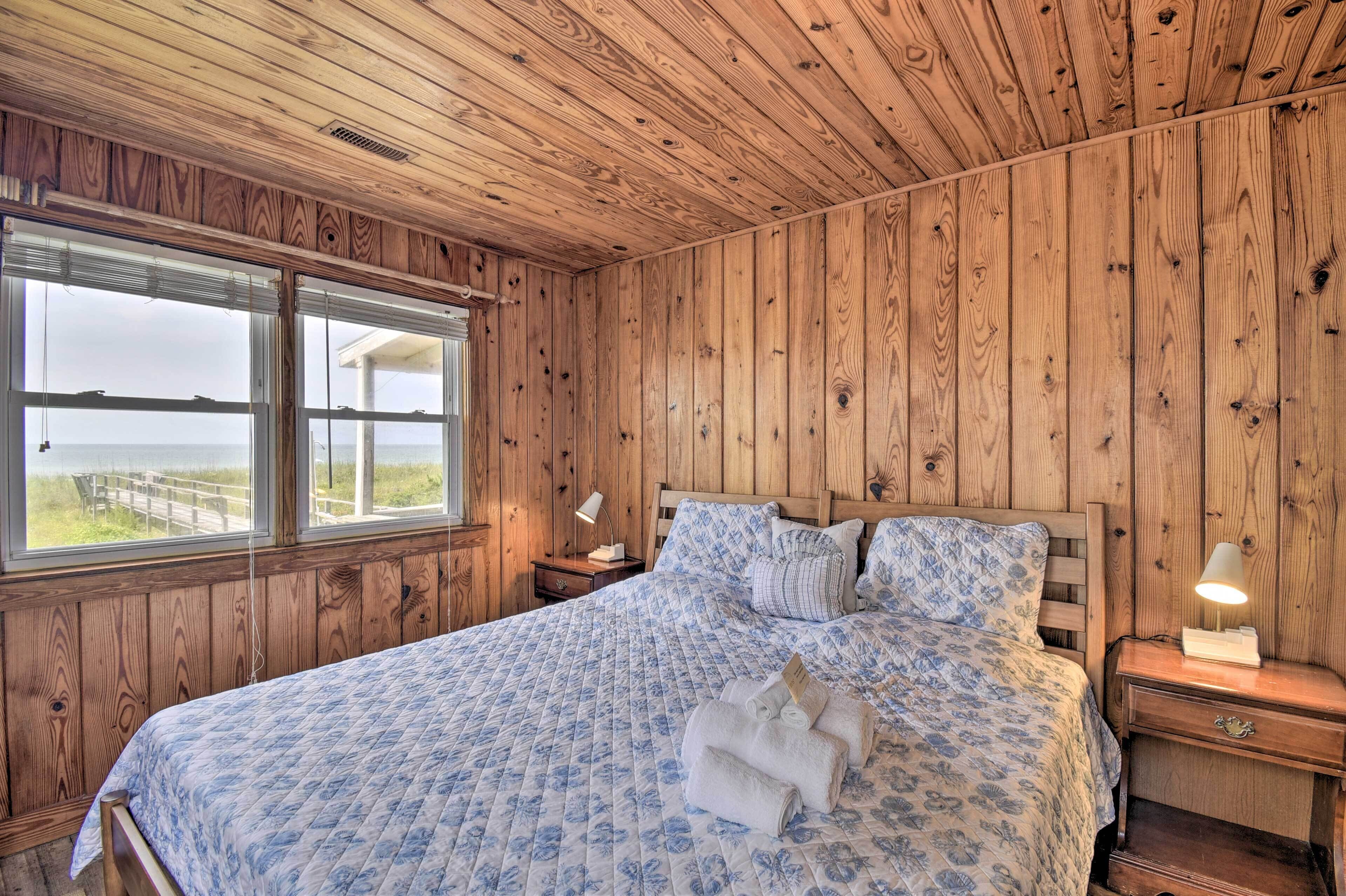 A wooden-paneled bedroom with a large bed, two bedside tables, and a view of the beach through a window.