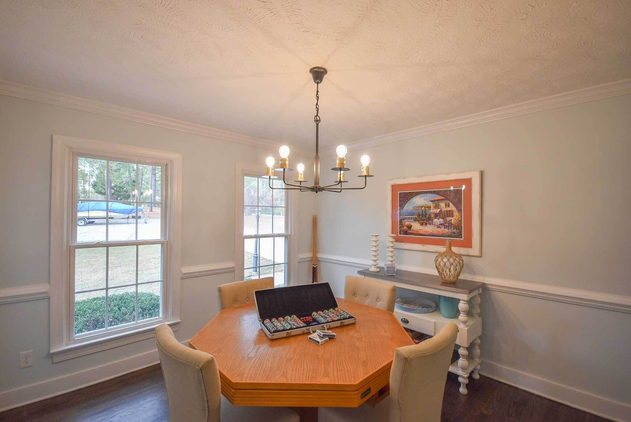 A dining room with a round wooden table, six chairs, a chandelier, a window with a view of a car, and a framed picture on the wall.