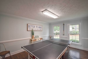 Una mesa de ping pong en una habitación con una lámpara en el techo, un cartel enmarcado en la pared y una ventana con vistas a la vegetación.