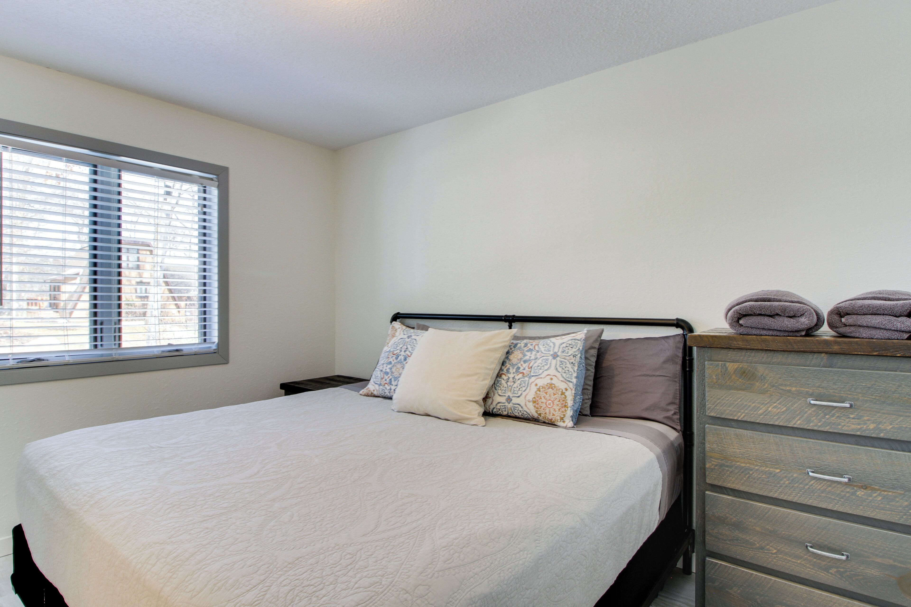 A neatly made bed with a white comforter and patterned pillows, a wooden dresser with drawers, and a window with blinds.