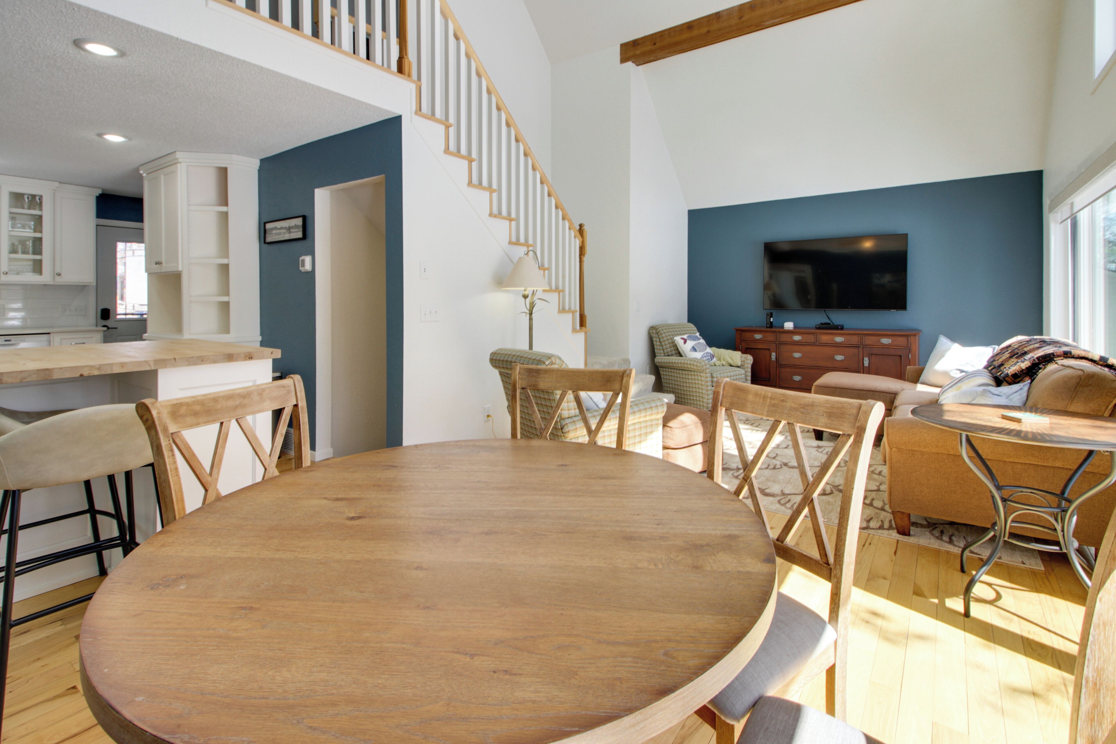 A spacious living area with a wooden dining table, comfortable seating, a staircase, a television, and a kitchen area.