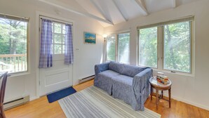 A room with a blue sofa, a small round table with books, and large windows with a view of trees. - Couples Getaway Near Beaches: Northport Cottage (Lincolnville)