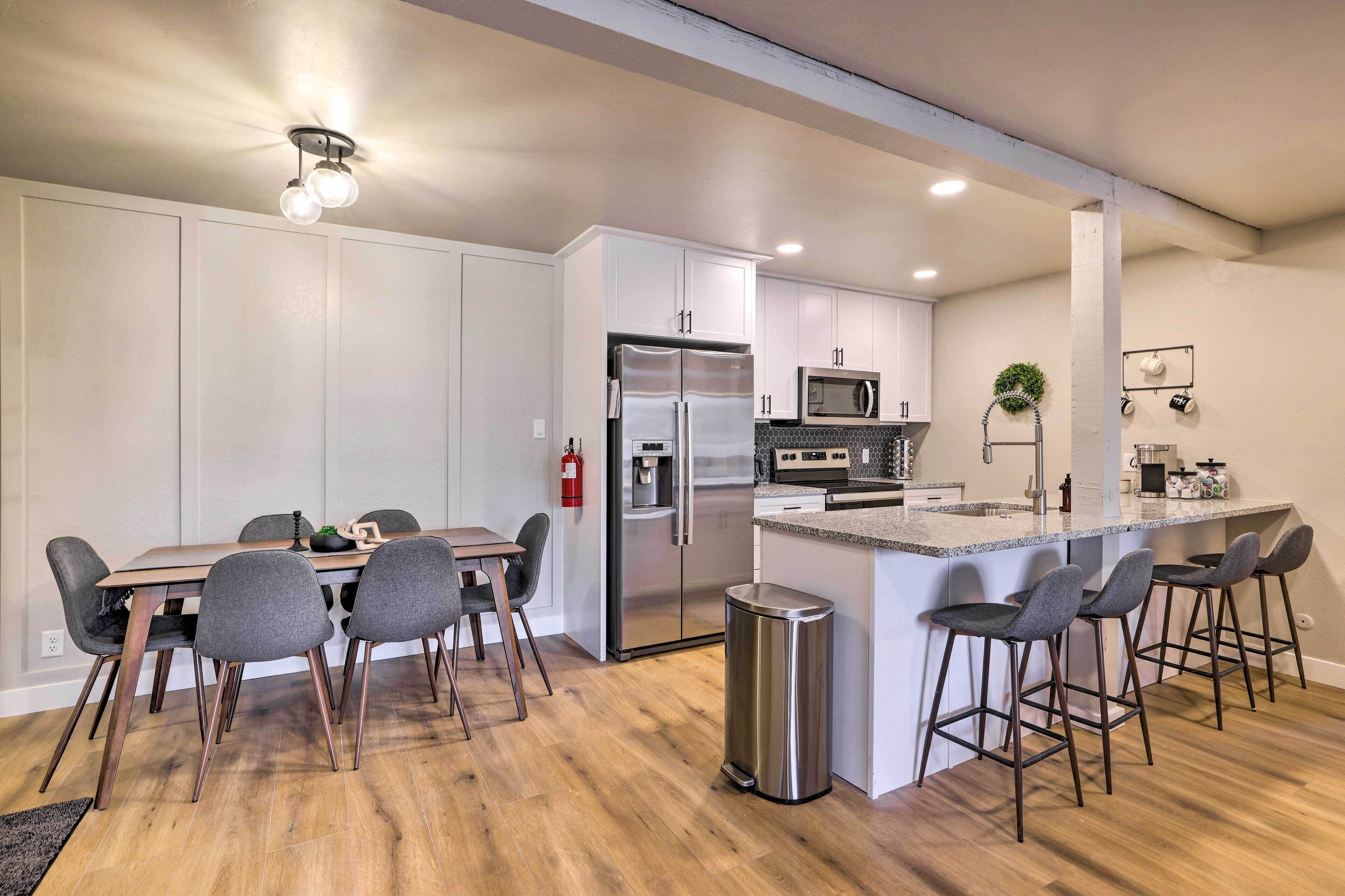 Una cocina moderna con armarios blancos, electrodomésticos de acero inoxidable, una zona de comedor con mesa de madera y sillas grises, y un pequeño bar con taburetes.