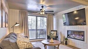 A living room with a sofa, a round coffee table, a fireplace, a ceiling fan, and a large window with curtains. - Brian Head Condo Near Lifts at Giant Steps! (Brian Head)