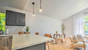 Una cuina moderna amb armaris foscos, una illa gran i il·luminàries penjants. Hi ha una zona de menjador amb cadires de fusta i una taula petita, i una zona d'estar amb un sofà i coixins.