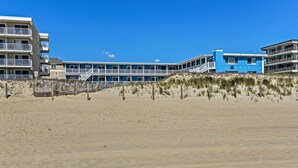 Un hotel frente a la playa con varios pisos, una playa arenosa en primer plano y un cielo azul despejado.