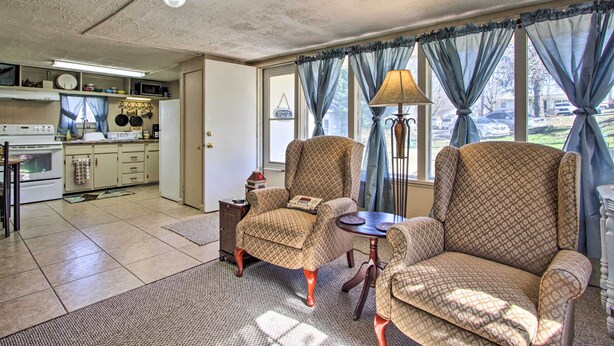A spacious living room with two patterned armchairs, a small table, and a lamp. A kitchen is visible in the background with a refrigerator, stove, and various kitchen items. - 2 Mi to Downtown Knoxville: Cozy Home w/ Patio (Knoxville)