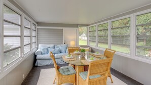 A sunroom with a glass table surrounded by wicker chairs, a blue sofa, and large windows offering a view of the outdoors. - Cozy Cottage in Historic Village: Near Cedar Point (Milan)