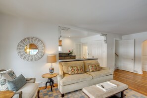 A living room with a sofa, a coffee table, and a side table with a lamp. There is a mirror on the wall and a door leading to another room.