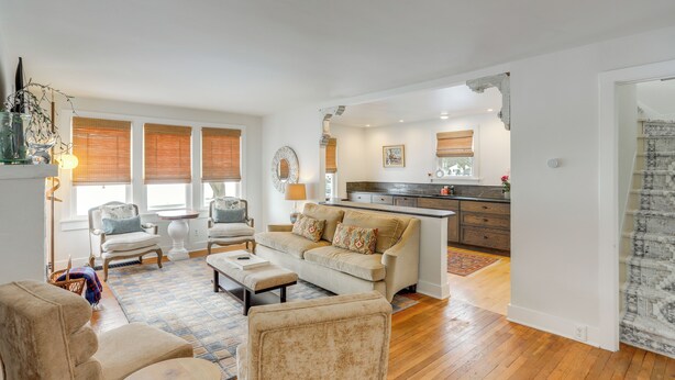 A spacious living room with a beige sofa, two armchairs, a coffee table, and a patterned rug. A kitchen with a dark countertop and wooden cabinets is visible in the background. - Cozy Cottage in Historic Village: Near Cedar Point (Milan)