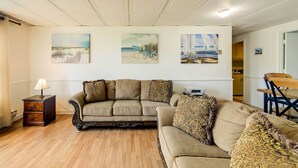 A living room with a sectional sofa, a wooden nightstand, and wall art depicting beach scenes. - 'hannah's Place' - Quaint & Cozy Apt by the Beach! (Emerald Isle)