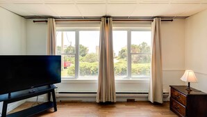 A room with a flat-screen TV on a black stand, a wooden dresser with a lamp, and a window with curtains. - 'hannah's Place' - Quaint & Cozy Apt by the Beach! (Emerald Isle)