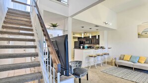 Un salon moderne avec un escalier, un fauteuil gris, un canapé avec des coussins jaunes et, au fond, un coin cuisine.