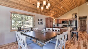 A spacious dining area with a large wooden table and white chairs, a wooden ceiling with exposed beams, and a view of the outdoors through large windows. - Fairplay Cabin w/ Wraparound Deck & Mountain Views (Fairplay)